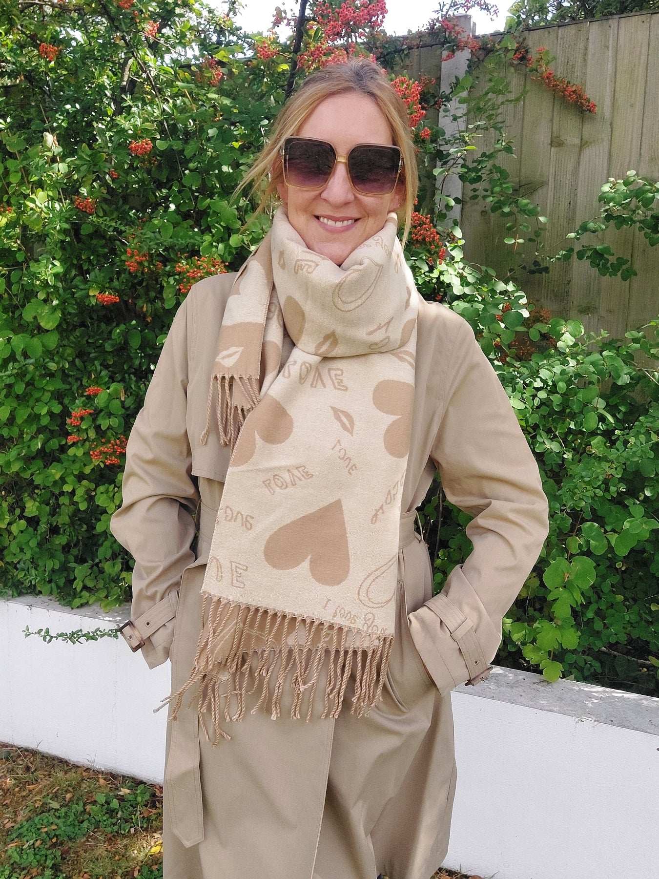 Woman wearing a beige coat and scarf with heart patterns, standing in front of greenery.