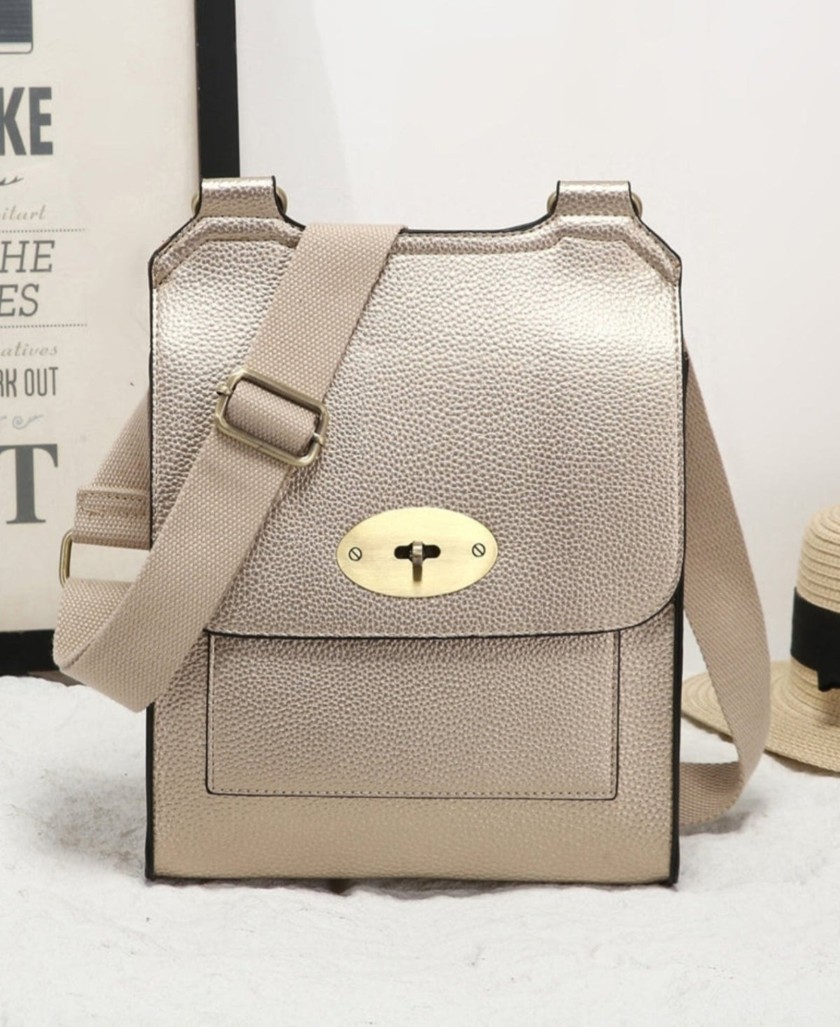 A metallic gold pebbled leather crossbody bag with a gold-tone turn-lock clasp and a beige fabric strap, displayed on a white textured surface next to a straw hat and a framed print.