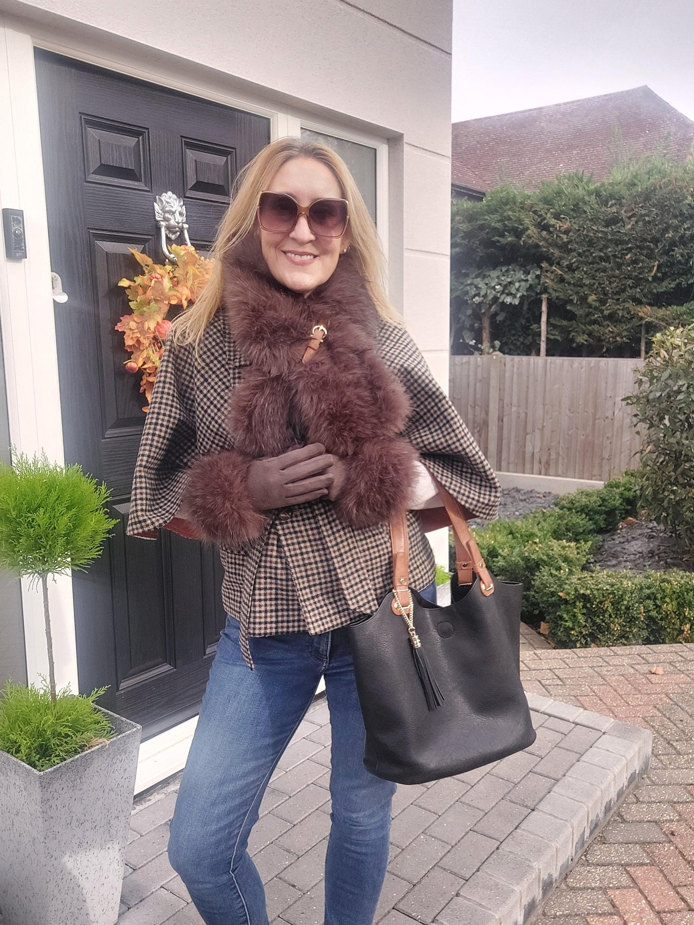 Person wearing a checkered coat with fur trim and carrying a black handbag outside a house.
