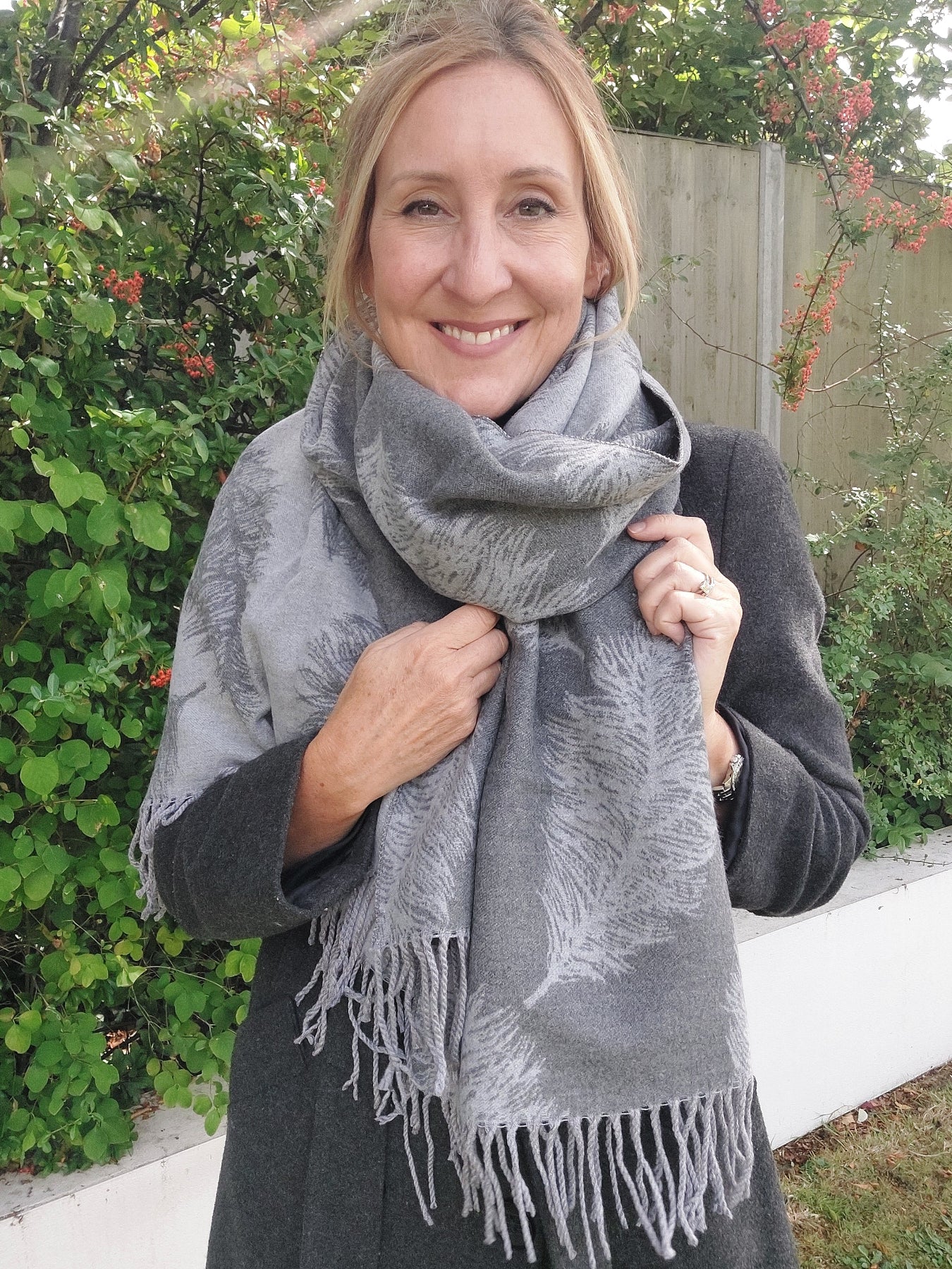 Woman wearing a gray scarf with fringe, standing outdoors with greenery in the background.