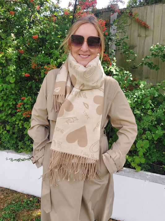 Woman wearing a beige coat and scarf with heart patterns, standing in front of greenery.